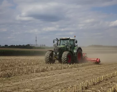 Un tracteur agricole vert avec des jantes rouges dans un champ labouré sous un ciel partiellement nuageux. Le tracteur est équipé d'un broyeur rouge attaché à l'arrière, soulevant de la poussière. Des rangées de chaumes de récolte s'étendent à l'infini, et des arbres et des lignes électriques sont visibles à l'horizon.