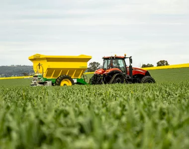 Un tracteur rouge tirant un grand épandeur jaune traverse un champ de cultures vertes, avec un paysage vallonné et des bandes de champs jaunes en arrière-plan sous un ciel nuageux.