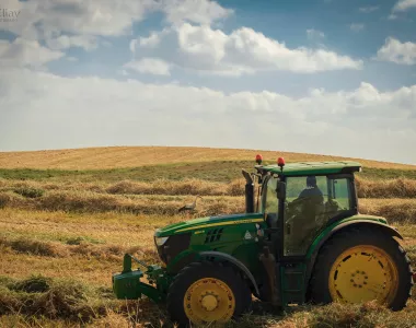 Tracteur agricole John Deere vert avec grandes roues jaunes, conduit dans un champ de chaume et d’herbes coupées, sous un ciel bleu partiellement nuageux, avec un oiseau marchant dans les sillons.